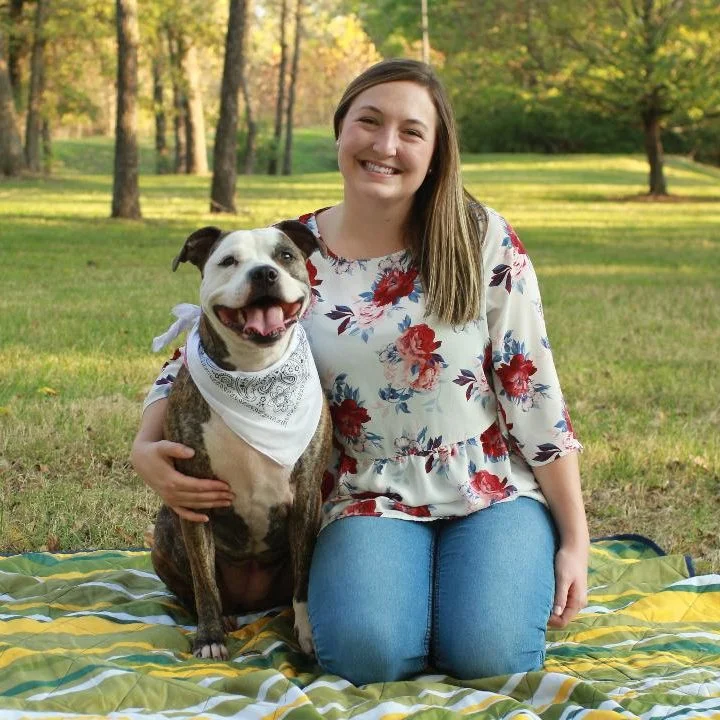 Dr. Laura Lorentzen sitting on a blanket outdoors, smiling next to a dog wearing a bandana.